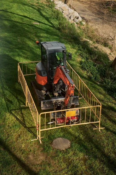 Mini excavator surrounded by fence above ground in a field or forest. Laying subway sewer pipes during the construction of a house. Digging trenches for a gas or oil pipeline. Earth moving, foundation