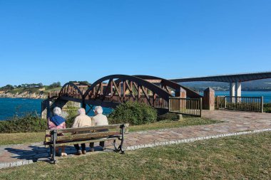 Tanımlanamayan yaşlı insanlar bir bankta oturup sağlarındaki Ribadeo nehri, Lugo, Galiçya, İspanya, Avrupa ve Azizler köprüsünün üzerindeki ahşap köprüyü izliyorlar..