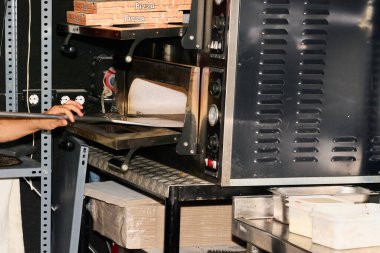 Tanımlanamayan pizzacı fırından pizza almaya hazırlanıyor. Fırının üstünde ve altında pizza kutuları monte edilmiş ve parçalanmış.