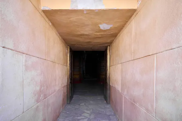 Narrow hallway with a low ceiling that leads to the entrance to a pyramid. The walls and ceiling are made of an orange-pink plaster. The floor is made of gray stone tiles