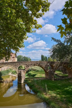 Meder nehri üzerindeki Queralt köprüsü, 11. yüzyıl, Vic, Osona, Barcelona, Katalonya, İspanya, sakin sular ve yanında bulutlar ve ağaç dalları ile mavi gökyüzü ve yeşil çayırlar..