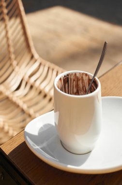 Üzerinde çikolata izleri olan beyaz bir fincan, güneş ışığıyla aydınlatılmış, örülmüş bir sandalyenin yanındaki ahşap bir masada..