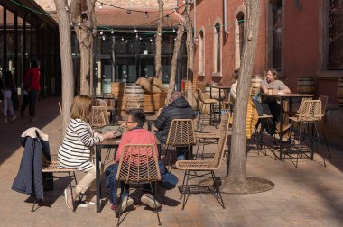 Barselona, İspanya - 06 Mart 2024: İnsanlar çıplak ağaçlar ve tarihi binalarla çevrili bir kafede güneşli bir günün tadını çıkarıyorlar.