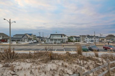 Modern evler neredeyse boş bir park alanı, ön planda bir ahşap çit ve bulutlu ama sakin gökyüzü sahnenin sakin atmosferine ekleniyor.