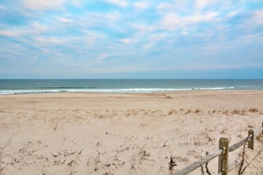 Kumlu bir patika, bulutlu bir gökyüzünün altında, ahşap bir çit ve kıyı bitkisiyle çevrili, sakin bir sahile doğru esiyor..