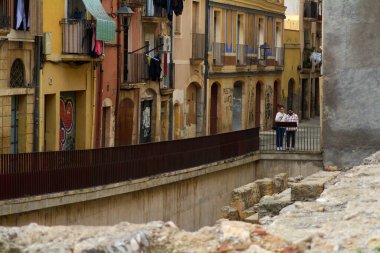 Tarragona, İspanya - 15 Nisan 2024: Tarragona 'da demir balkonları ve yıpranmış duvarları olan eski binaların yanında yürüyen bir çift, tarihin ve çağdaş yaşamın büyüleyici bir karışımını gösteriyor. Asılmış giysiler insan eli değmesine neden olur..