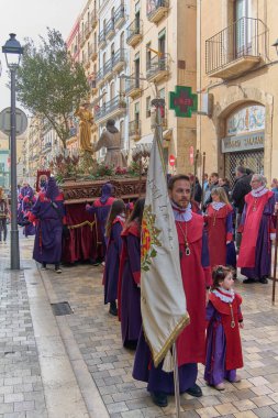 Tarragona, İspanya - 23 Nisan 2024: Eski binalar ve seyircilerle çevrili bir kaldırım taşı caddesinde mor cüppeli tövbekçilerle geleneksel geçit töreni
