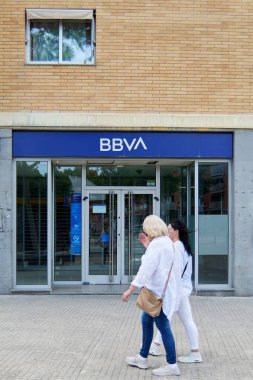 Viladecanlar, Barselona, İspanya-26 Mayıs 2024: BBVA şubesinin ana girişinin cam kapılar ve bir ATM ile görüntüsü. İki kadın bankanın önünden geçiyor..