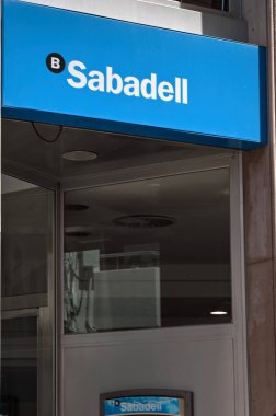 Sitges, Barcelona, İspanya-Haziran 3, 2024: Bir Banco Sabadell girişinin ikonik mavi ve beyaz tabelasını gün ışığı altında karşısındaki bir binayı yansıtan görüntüsü.