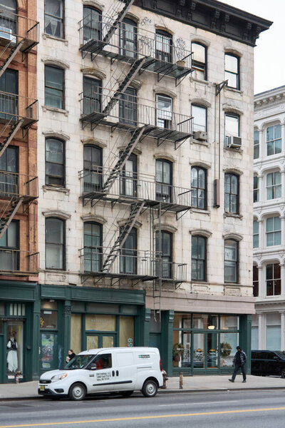 New York, United States - August 27, 2024: Old building with fire escapes in a historic neighborhood of New York. A parked vehicle and a pedestrian add life to the urban scene.