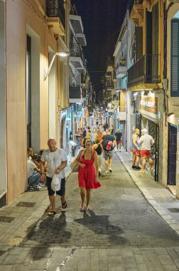 Sitges, Barcelona-Eylül 04.2024: İnsanlar, balkonlu binalarla çevrili, sicim lambalarıyla aydınlatılmış bir sokakta yürüyor.