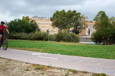 Viladecanlar, Barselona-Septembre 10.2024: Binalar ve doğa ile çevrili, kentsel ve spor temaları için mükemmel bir yolda yürüyen bir bisikletçinin görüntüsü. Sürdürülebilir hareketlilik ve açık hava egzersizleri için ideal.