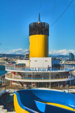 Barselona 'da. Espain - 20 Ekim 2024: Barcelona limanındaki Costa Fortuna yolcu gemisinin arkasından kentsel ufuk çizgisi ve deniz manzarası. Deniz taşımacılığı ve seyahat turizmi konuları için ideal.