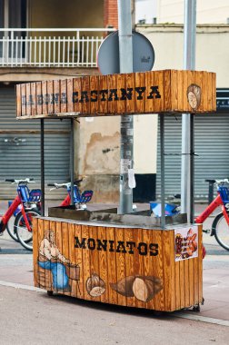 Viladecans. Spain - November 03,2024: Traditional wooden cart for selling chestnuts and sweet potatoes, positioned on an urban street with bicycles nearby, representing local street food vendors. clipart