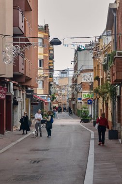 Viladecanlar. İspanya - Kasım 03.2024: Viladecan 'daki yaya sokağı dekoratif ışıkları ve çevresi konut binaları ve yerel dükkanlarla çevrili gezen insanlar, rahat bir kentsel atmosfer yaratıyor.