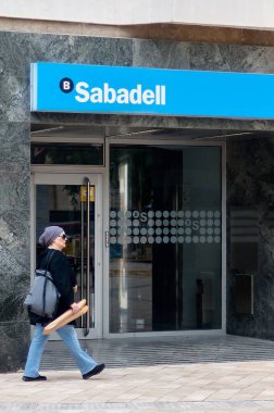 Viladecanlar. İspanya - 7 Kasım 2024: Baget taşıyan bir kadın Sabadell 'deki bir banka şubesinin önünden geçerek modern cephe ve cam kapılar sergiledi.