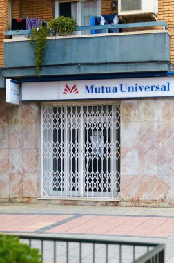 Viladecans. Spain - November 07,2024: The image shows the closed storefront of a Mutua Universal office with a security gate. The sign above the entrance displays the company name and logo, indicating a professional setting. clipart