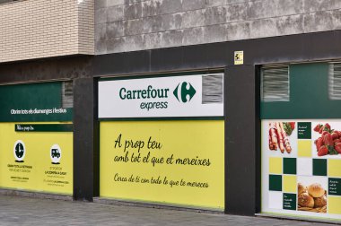 Viladecanlar. İspanya - 7 Kasım 2024: Bir Carrefour Express mağazasının dışı, günlük açılış saatlerini ve ev teslimat hizmetlerini ön plana çıkaran reklamlar. Metinler Katalanca ve İspanyolca.