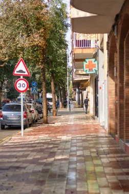 Viladecanlar. İspanya - 9 Kasım 2024: Viladekan kentsel sokak manzarası üzerinde bir eczane işareti, hız sınırı işareti ve kaldırımda yürüyen yayalar.