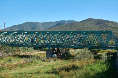 Montcada i Reixac. İspanya - 12 Kasım 2024: Ripoll Nehri 'ni geçen yeşil çelik köprünün yeşil bitki örtüsü ve açık mavi gökyüzü altındaki yuvarlanan tepeler tarafından çerçevelenmiş manzaralı bir manzarası.