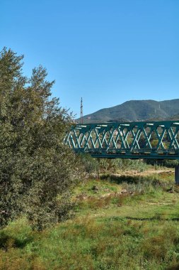 Montcada i Reixac. Spain - November 12,2024: A green steel truss bridge spans the Ripoll river, set against a backdrop of verdant hills and a clear sky, creating a tranquil and scenic landscape. clipart