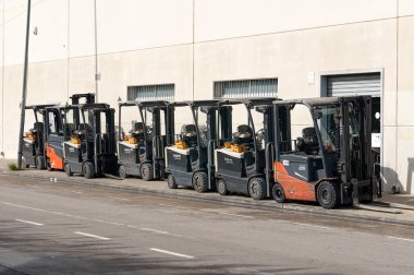 Viladecanlar. İspanya - 16 Kasım 2024: Kaldırıma düzgün bir sıra halinde park etmiş bir grup forklift. Sahne, şehir ortamında endüstriyel ekipmanların organizasyonunu ve hazırlık durumunu yakalıyor..