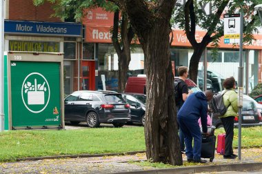 Viladecans. Spain - November 18,2024: Group of people waiting at an urban bus stop next to a parked Mercadona delivery truck. The scene captures the daily routine in the city, highlighting public transport and commerce. clipart