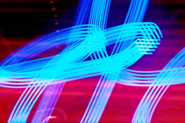 Mavi ışık yolları kırmızı ve koyu arkaplan karşısında kıvrımlı ve döngülü şekiller oluşturarak bu uzun pozlu çekimde dinamik ve soyut bir görsel deneyim sunar..