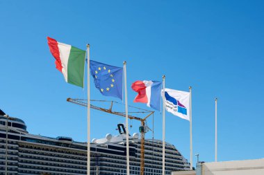 Marseille. France  - December 02, 2024: Shows flags of Italy, the European Union, France, and a cruise line logo flying at a port, with a large cruise ship and construction crane in the background. clipart