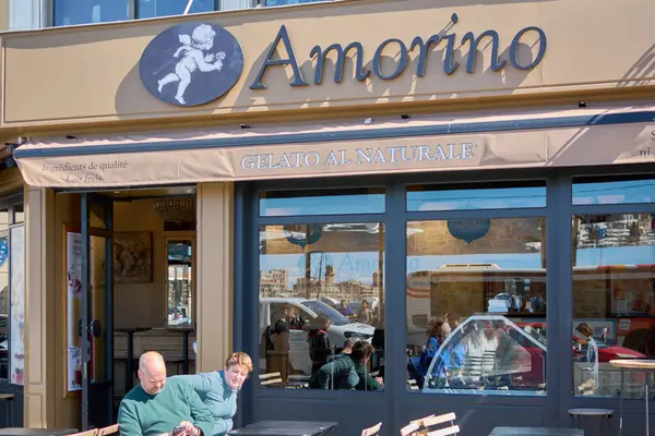 Marsilya, Fransa, 4 Aralık 2024 Amorino gelato dükkanında güneşli bir gün. Müşteriler dışarıda yemek yemeyi severler. Mağaza vitrininde Amorino logosu ve Gelato al Naturale görünüyor, rahat bir atmosfer yaratıyor..