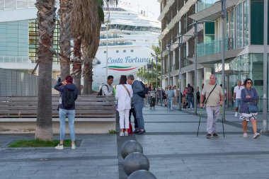 Savona. Italia  - December 07, 2024: Tourists walking and taking photos near the Costa Fortuna cruise ship docked at the port of Savona, Italy, with modern buildings and palm trees in the background. clipart