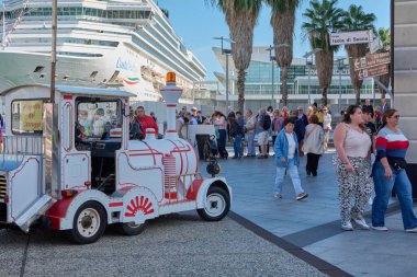 Savona. İtalya - 07 Aralık 2024: Savona limanında turist treni ve arka planda çok sayıda insanın yürüyüş yaptığı ve güneşli günün tadını çıkardığı hareketli bir gezi sahnesi.