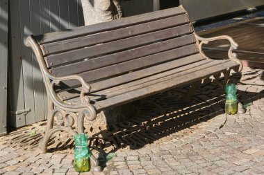 Her iki ucunda kül tablası olarak kullanılan geri dönüşümlü yeşil plastik şişelerin yer aldığı bir kaldırım taşı caddesinde işlenmiş demir yapıya sahip ahşap bir bank. Bankta sakin bir kent alanı var..