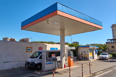 Civitavecchia. İtalya - 17 Aralık 2024: Beyaz minibüs birden fazla pompa ve kırmızı ve beyaz direklerle şehir benzin istasyonunda yakıt ikmali yapıyor. Arka planda işaret ve yangın söndürücü var. Kavram: Kentsel yakıt ikmali.