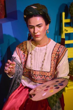 Madrid, Spain - January 14, 2025: Frida Kahlo in traditional attire, holding a paint palette and a paintbrush, sitting in a cultural setting with a decorative background. clipart