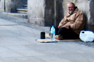 Madrid, İspanya - 20 Ocak 2025: Kahverengi ceketli evsiz bir adam duvardaki bir karton üzerinde oturmuş, elinde bir şişe su ve bir bardak tutuyordu. Sahne Madrid 'deki evsizlerin sorununu ortaya çıkarıyor..