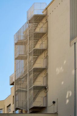 Bej bir binanın yan tarafına eklenmiş dış metal yangın çıkışı. Yapı dikey metal çubuklarla çevrili ve çeşitli katlara yayılarak kentsel güvenlik özelliklerini yansıtıyor.