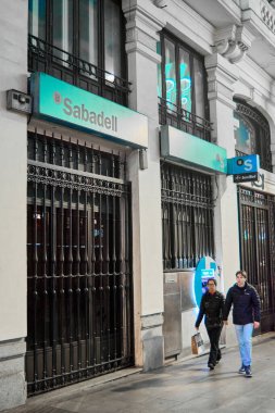 Madrid mi? İspanya - 24 Ocak 2025 Sabadell Bank 'ın gece manzarası aydınlatma işareti, ATM' si ve yakınlarda yürüyen iki kişi. Kavram: Kentsel bankacılık simgeleri