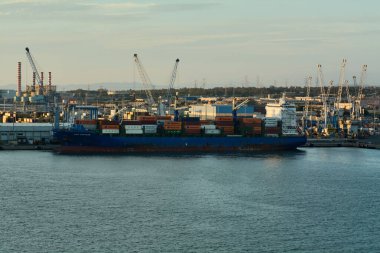 Civitavecchia. İtalya - Şubat 06, 2025: Renkli konteynırlarla dolu ve turna ve sanayi binalarıyla çevrili yoğun bir sanayi limanına yanaşan büyük bir kargo gemisi.