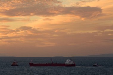 Büyük kırmızı bir kargo gemisine, güzel bir gün batımında sakin bir denizde, ufukta görünen uzak dağlarla birlikte bir römorkör eşlik eder..