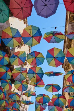 Parlak, çok renkli şemsiyeler açık mavi gökyüzüne karşı neşeli bir tepe örtüsü oluşturarak neşe ve yaratıcılığı çağrıştırıyor. Kavram: kaprisli açık hava sanatı