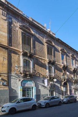 Catania 'da. İtalya - 03 Mart 2025: Yaşlanmış cepheli tarihi taş bina, süslü demir balkonlar ve canlı şehir yaşamında berrak mavi gökyüzünün altına kurulmuş şehir cazibesi.