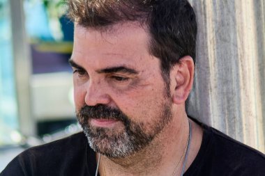 Kısa siyah saçlı, siyah tişörtlü ve altın kolyeli, açık havada zarif, bulanık arka planlı bir adamın modern portresi..