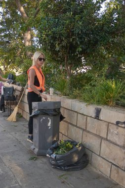 Girit. - 27 Mart 2025: Turuncu güvenlik yeleği giyen şehir temizlikçisi, bir çöp tenekesinin yanında duruyor ve içi gazete kupürleriyle dolu bir çöp torbası ile yakınlardaki bir duvara yaslanmış bir süpürge..