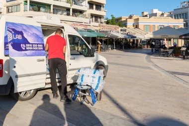 Girit. Yunanistan - Nisan 02, 2025: Yoğun bir meydanda, restoranlar ve kafelerle dolu beyaz bir minibüste buz dağıtım işçisi, kentsel tedarik zincirlerinin günlük faaliyetlerini vurguluyor.