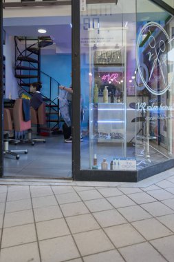 Girit - Yunanistan - Nisan 03, 2025: Parlak sandalyeli, spiral merdivenli, ışıklı saç bakım raflı ve makas logosu ve neon ışıklı cam kapılı modern salon.