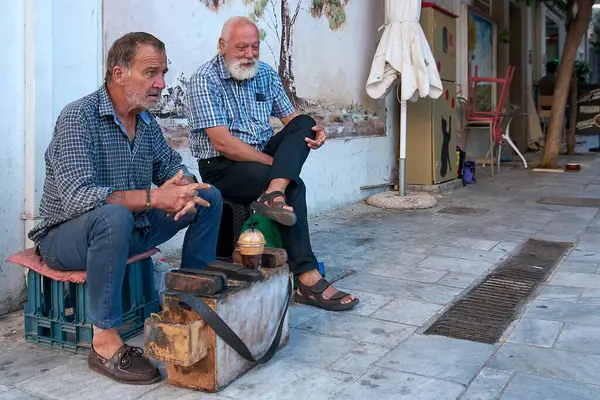Girit. Yunanistan - Nisan 02, 2025: Canlı bir Yunan şehir atmosferinde geleneksel bir ticareti yansıtan, fırça ve cilalı bir sandığın üzerinde oturan ayakkabı parlatıcısı.