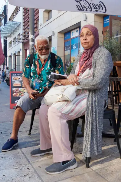 Girit. Yunanistan - Nisan 07, 2025: Bir sokak kafede oturan yaşlı bir çift canlı sohbete daldı. Sahnede canlı işaretler, bereketli yeşillik ve güneşli kentsel çevre yer alıyor..