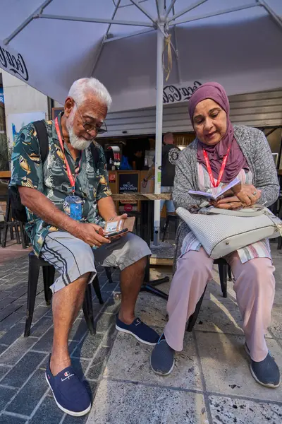 Girit. Yunanistan - Nisan 07, 2025: Açık bir kafede dinlenen iki yaşlı turist, günlük tatil kıyafetli bir adam ve güneşli bir öğleden sonra şehir ortamında eğlenen tesettür içindeki bir kadın.