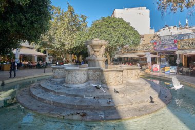 Girit. Yunanistan - Nisan 07, 2025: Muhteşem Morosini çeşmesi, Heraklions tarihi meydanında canlı yeşillikler ve hareketli aktivitelerle çevrelenmiş, su püskürten dört karmaşık oyulmuş aslanın yer aldığı.